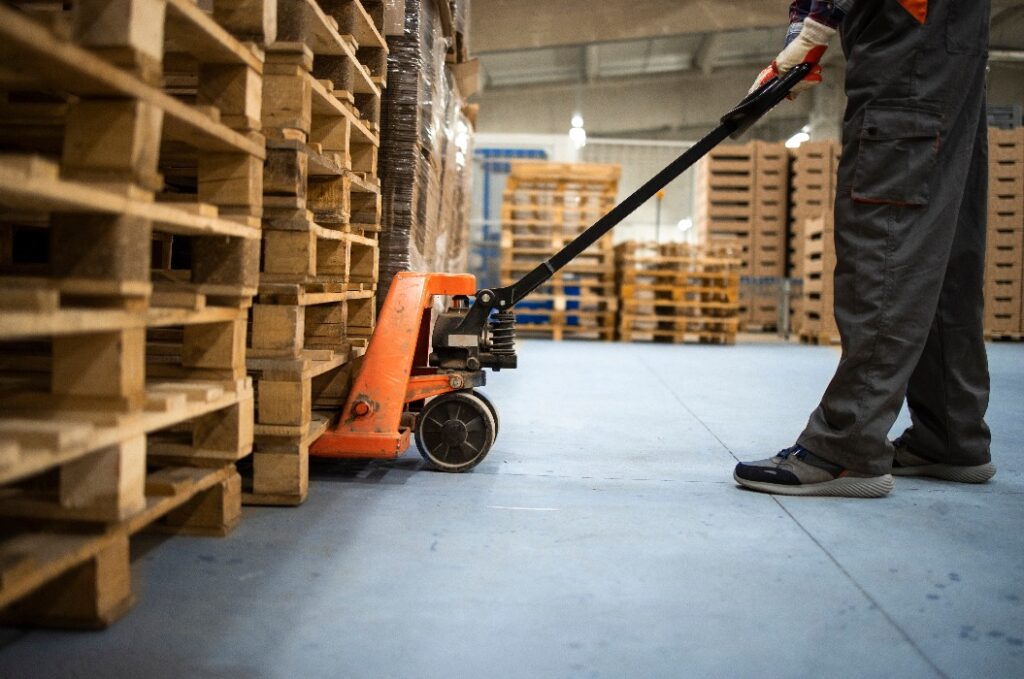 Arbeiten im Lagerhaus. Niedriger Blickwinkel auf einen nicht erkennbaren Arbeiter, der eine Palette mit einem manuellen Gabelstapler anhebt.