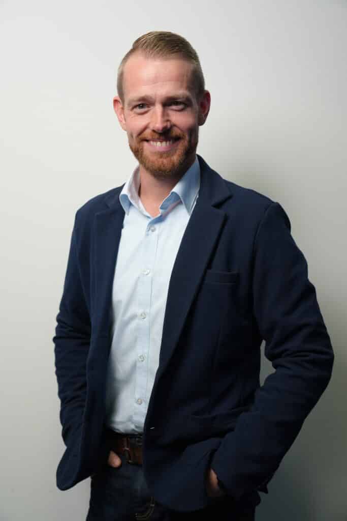 Ein lächelnder Mann mit Bart in blauem Sakko und heller Hose vor hellem Hintergrund